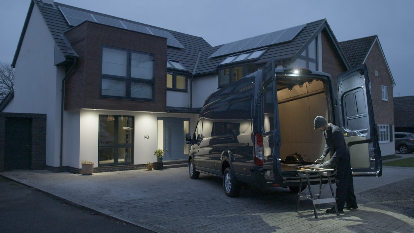 Ford Transit in Blau. Heckansicht, parkend vor einem Haus bei Nacht. Mann demonstriert die LED-Außenleuchte.
