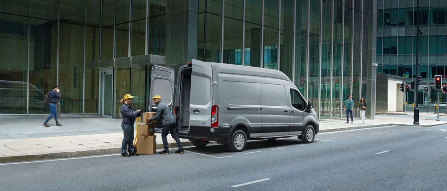 Ford E-Transit in Silber. Ansicht der geöffneten Hecktür, Frau und Mann beladen das Fahrzeug mit Kisten