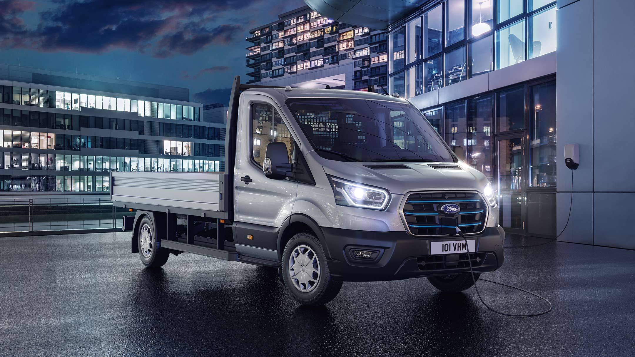 Ford E-Transit Fahrgestell in Silber. Dreiviertelansicht, stehend auf einem Parkplatz bei Nacht. 