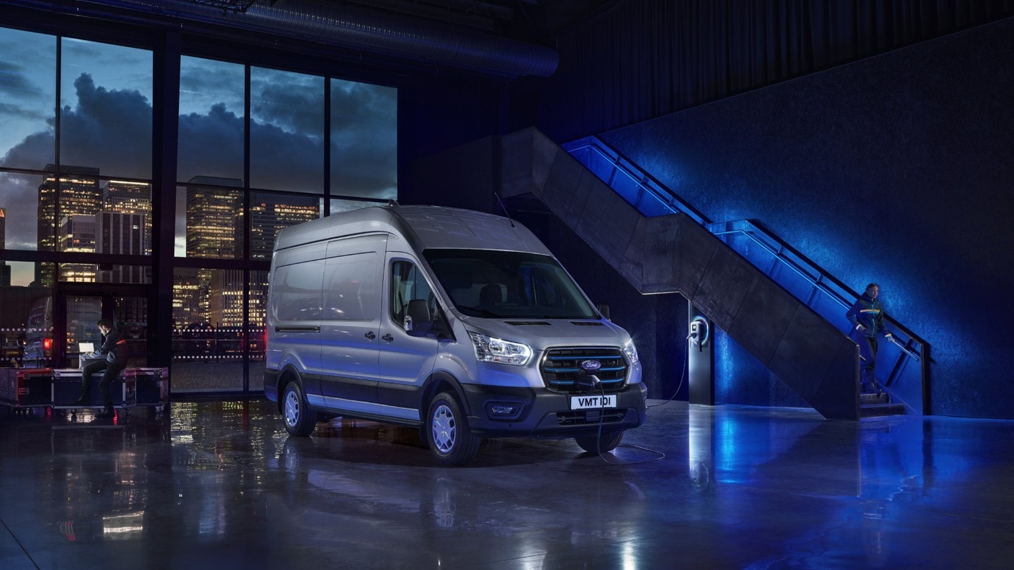 Ford E-Transit in Silber. Dreiviertelansicht, parkt in Lagerhalle mit blauer Ambientebeleuchtung
