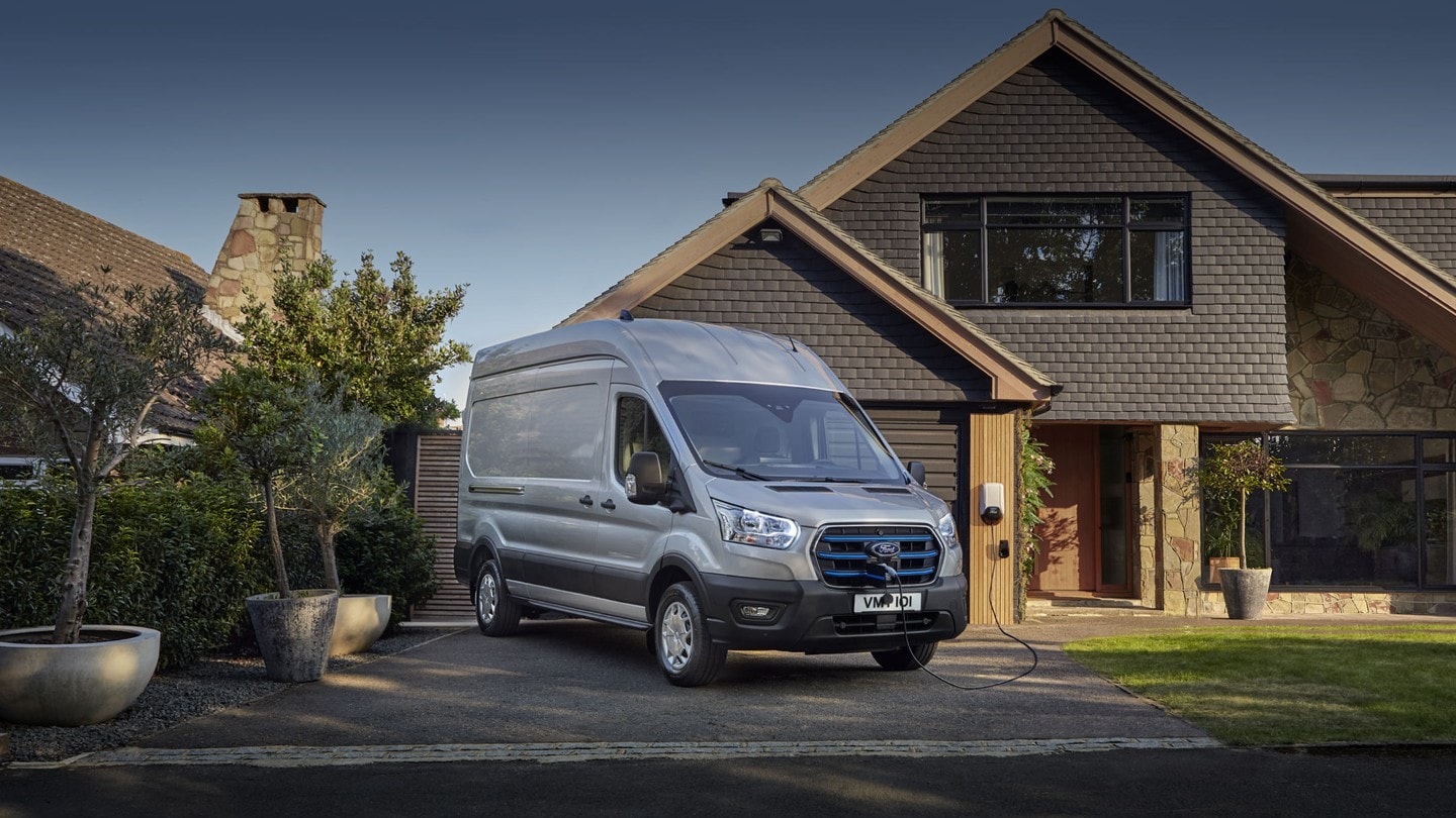 Ford E-Transit in Silber. Dreiviertelansicht, parkend vor einem Einfamilienhaus.