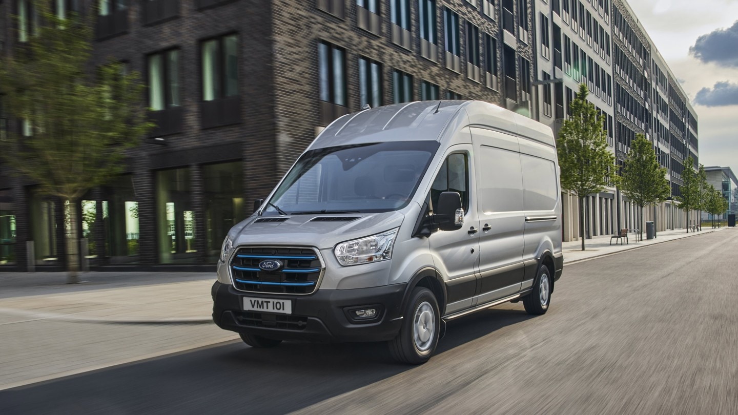 Ford E-Transit Kastenwagen in Silber. Dreiviertelansicht von vorne, fahrend entlang Hochhäusern