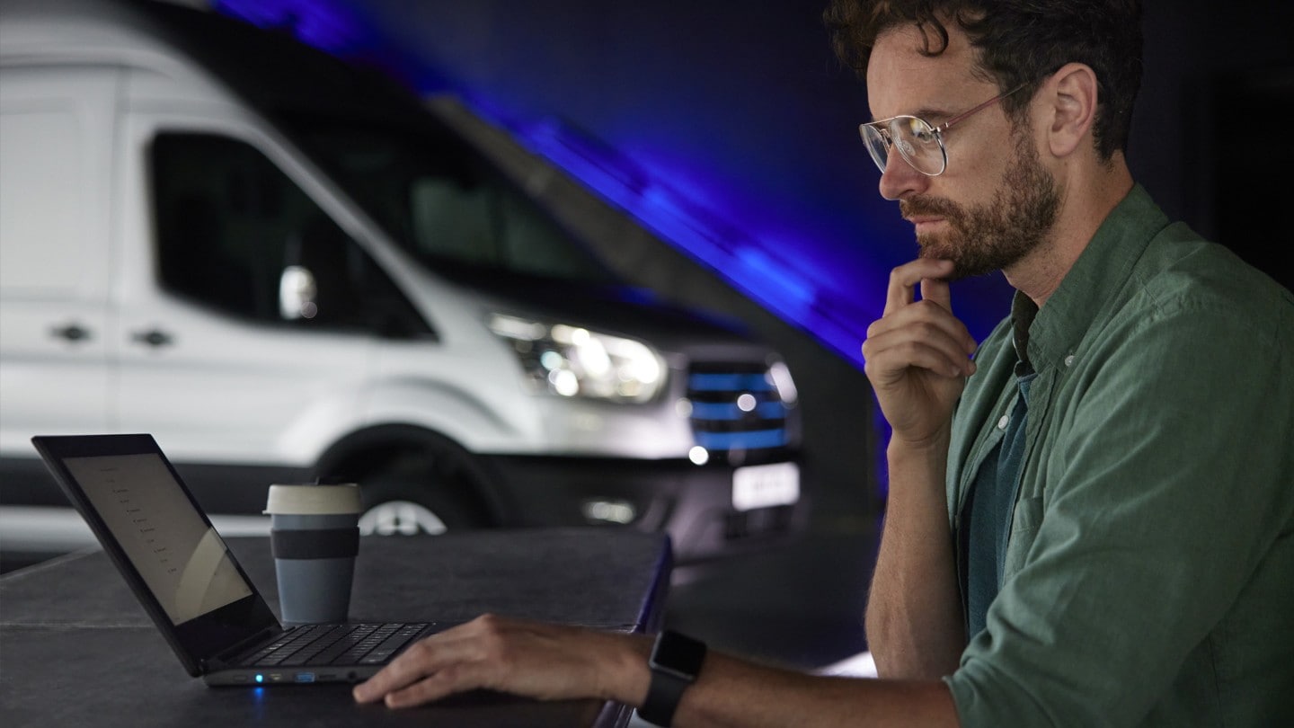 Mann sitzt vor Laptop. Im Hintergrund steht ein Ford E-Transit in Silber.