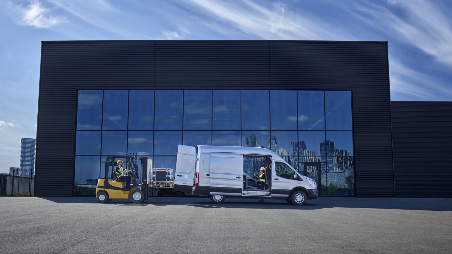 Ford E-Transit Kastenwagen in Silber. Seitenansicht, parkend vor modernem Gebäude mit geöffneter Schiebetür und Hecktür