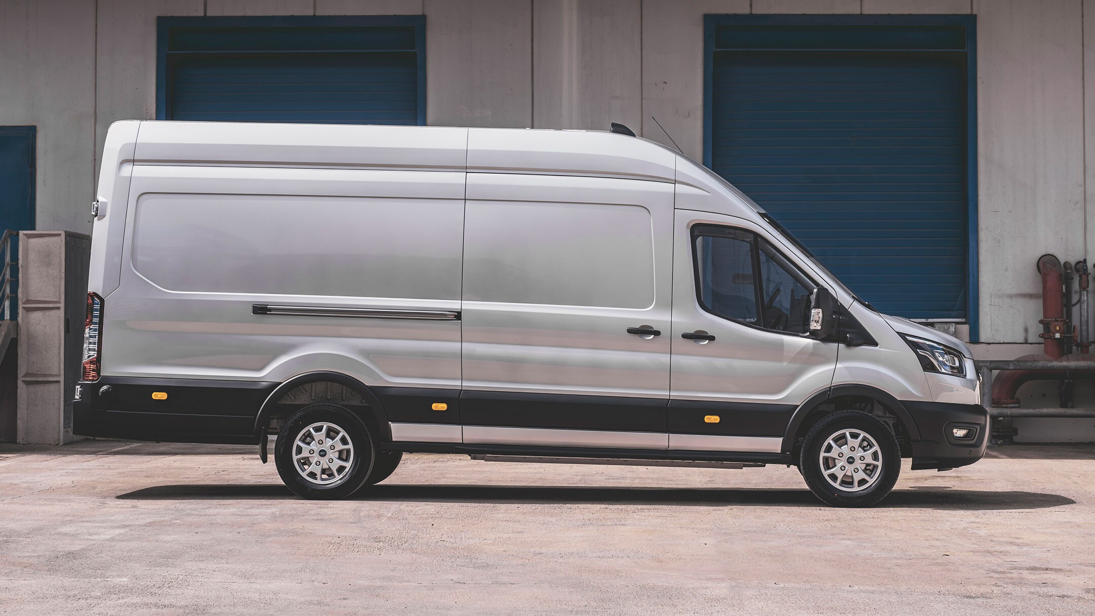 Ford E-Transit in Silber. Seitenansicht, stehend vor einer Lagerhalle.