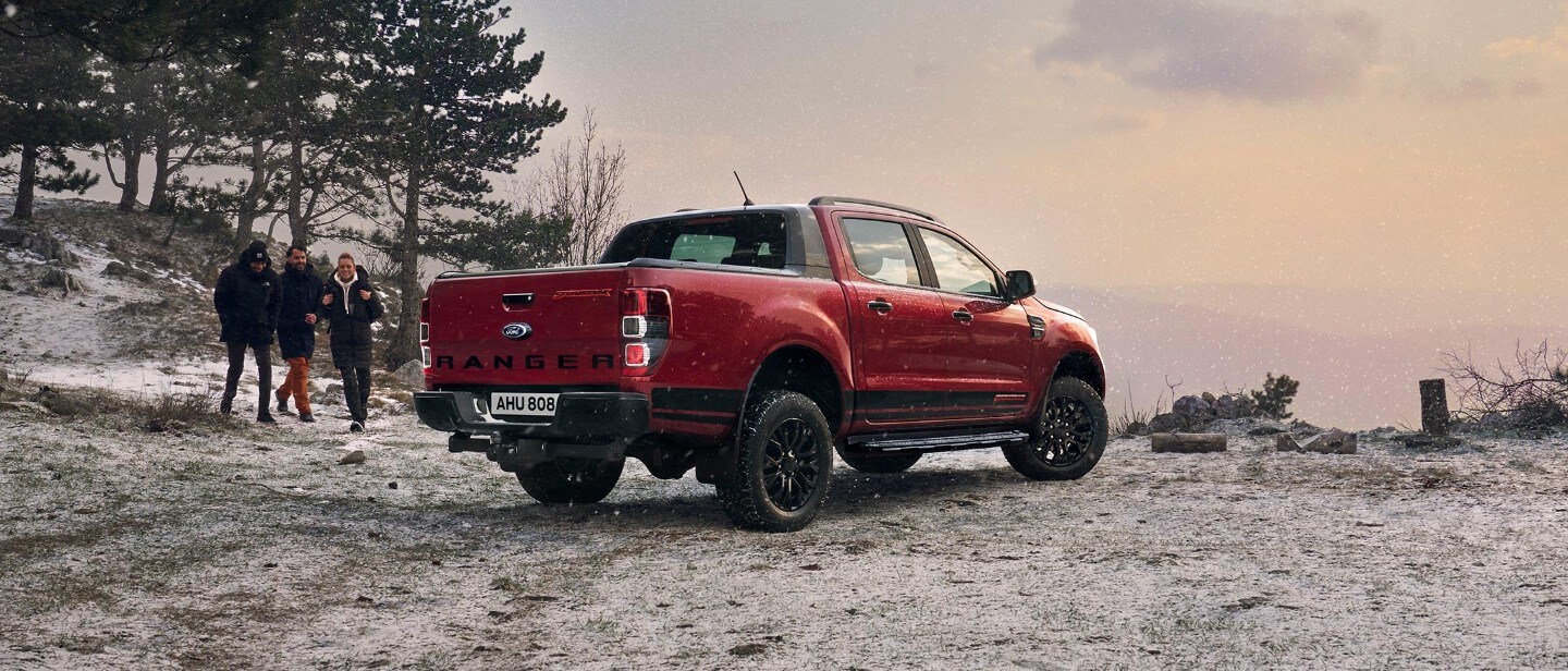 Ford Ranger Stormtrak in Rot. Dreiviertelansicht von hinten, parkend auf verschneitem, bergigem Untergrund und drei Personen sie auf das Fahrzeug zugehen
