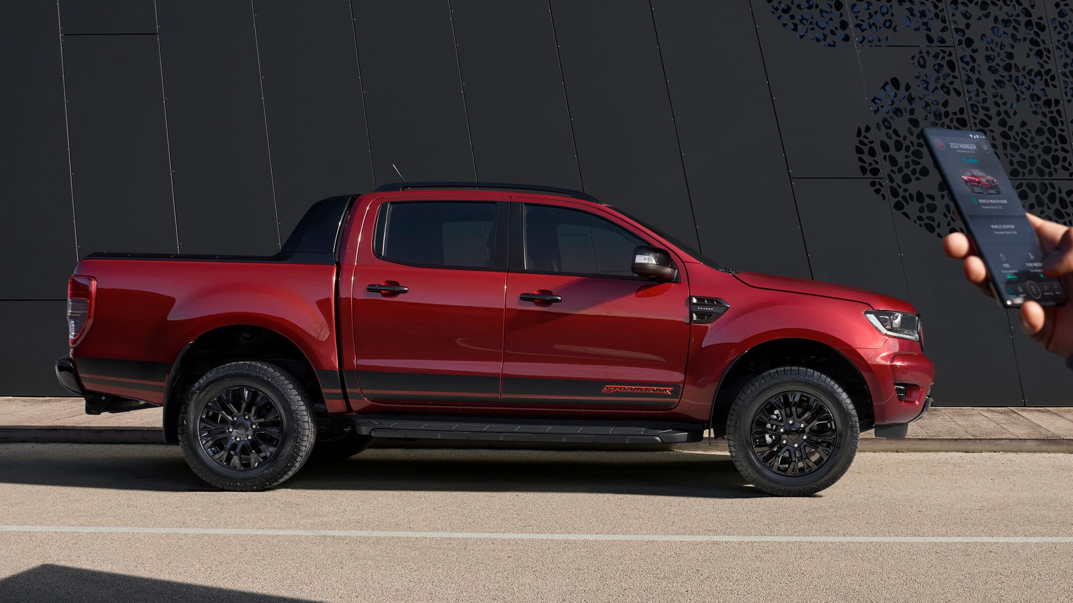 Ford Ranger Stormtrak in Rot. Seitenansicht, parkend am Straßenrand und Hand, die ein Mobiltelefon hält