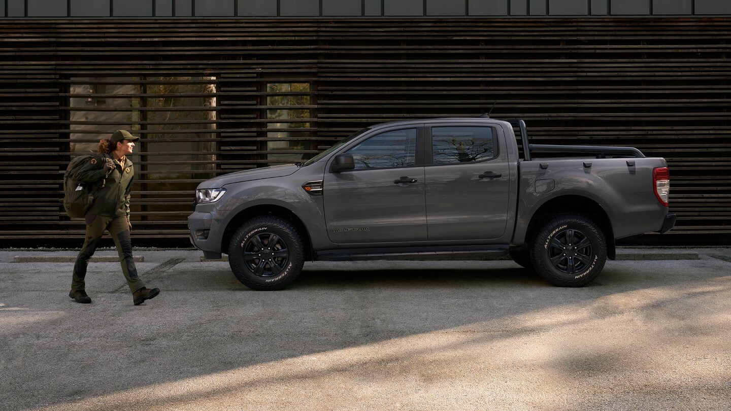 Ford Ranger Wolftrak in Grau. Seitenansicht, parkend neben einem modernen Gebäude und Frau in Outdoor-Kleidung mit Gepäck