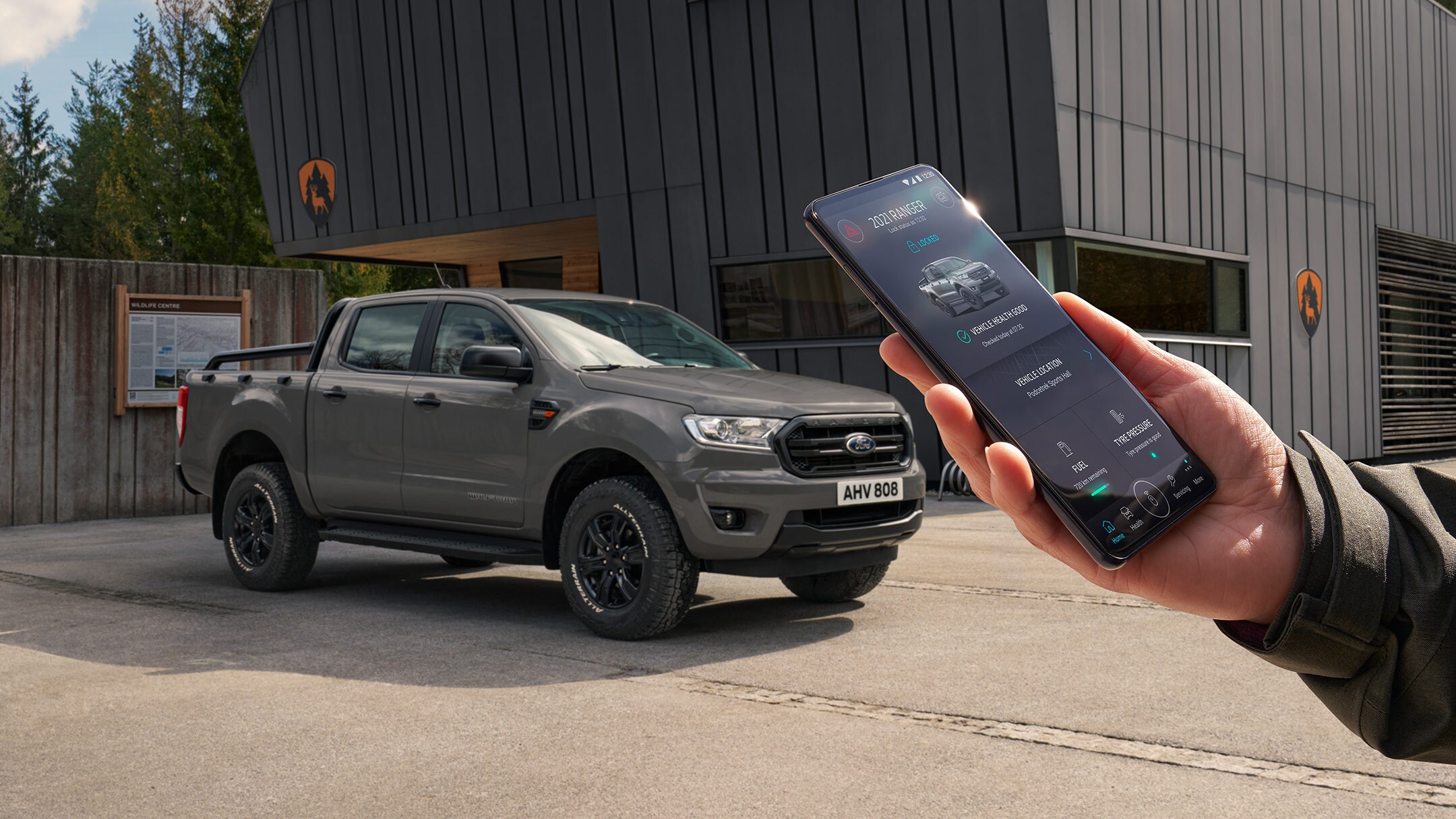 Ford Ranger Wolftrak in Grau. Dreiviertelansicht, parkend vor einem modernen Gebäude und Hand, die ein Mobiltelefon hält