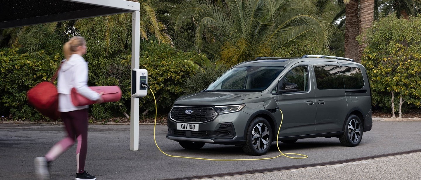 Ford Tourneo Connect in Grau. Dreiviertelansicht, an einer Ladestation angeschlossen. Frau geht zum Fahrzeug.