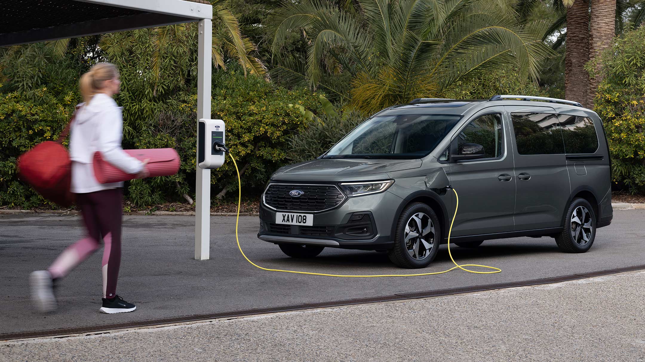 Ford Tourneo Connect in Grau. Dreiviertelansicht, an einer Ladestation angeschlossen. Frau geht zum Fahrzeug.