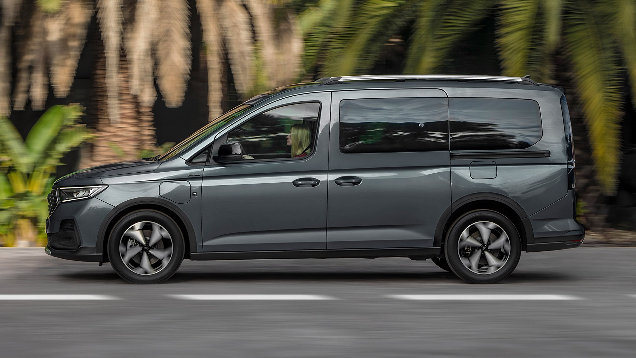 Ford Tourneo Connect in Grau. Seitenansicht, fahrend auf einer Straße. Frau sitzt im Fahrersitz.