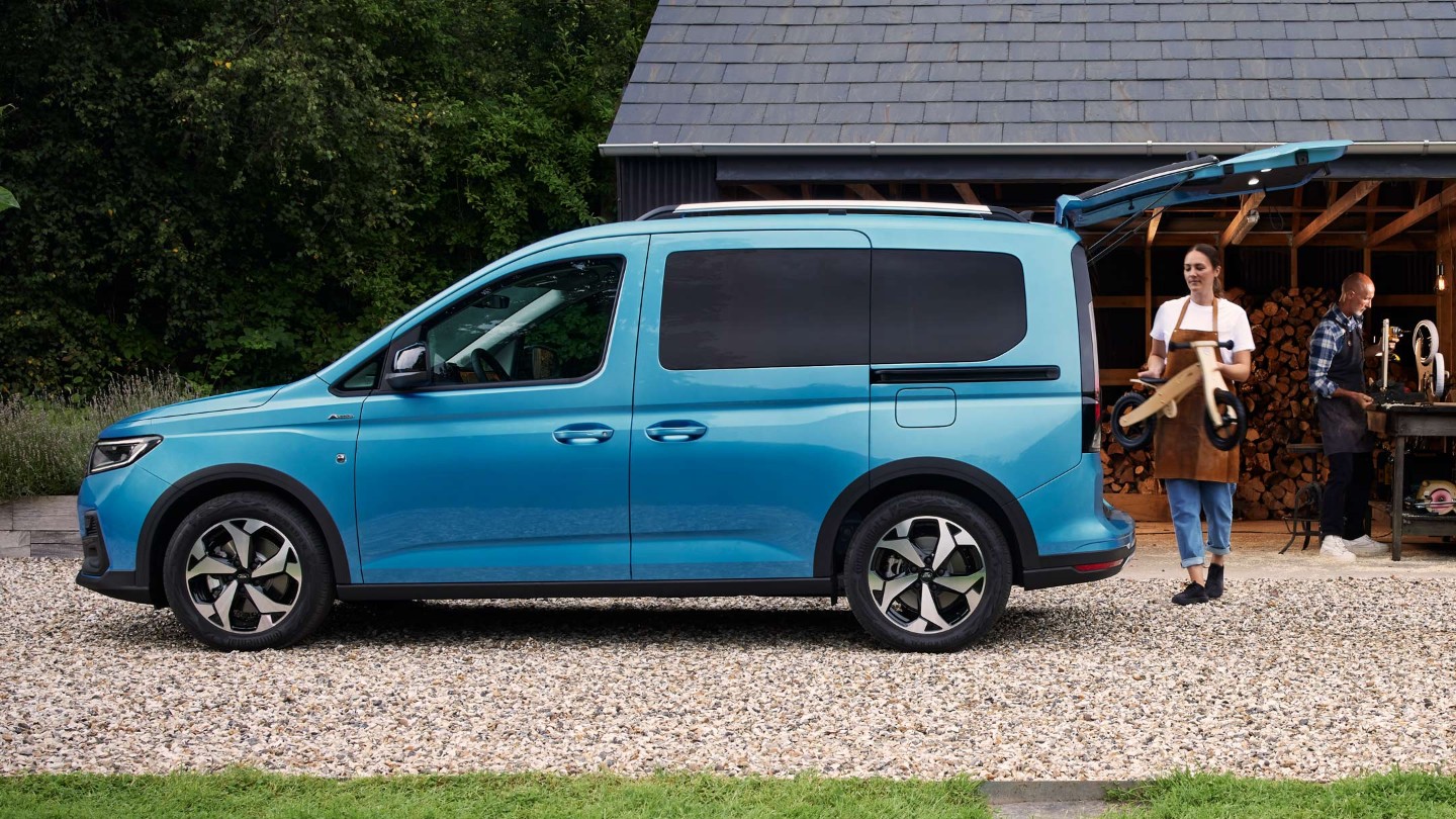 Ford Tourneo Connect in Boundless Blue. Seitenansicht mit geöffneter Hecktür, Frau belädt Gepäckraum