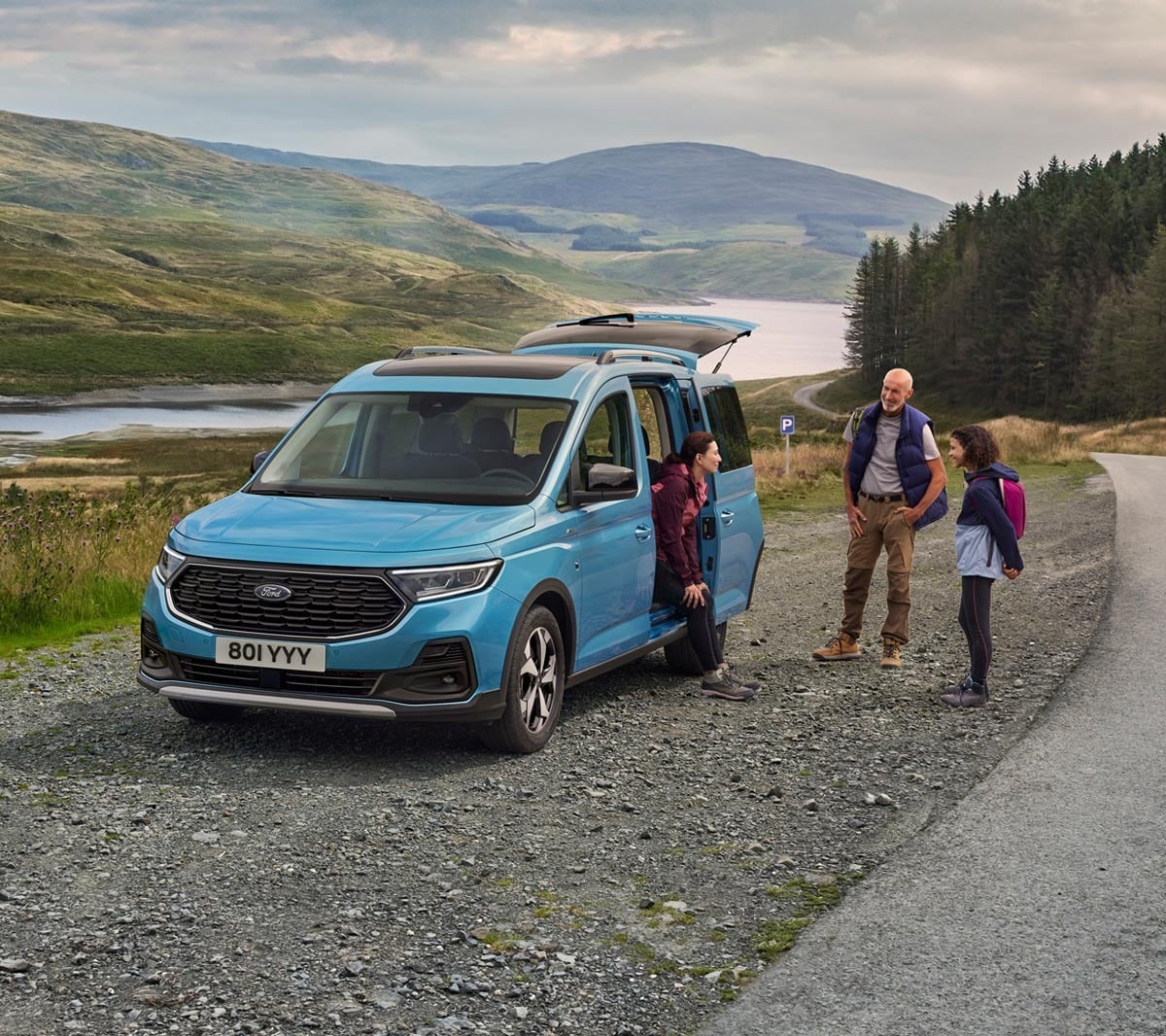 Ford Tourneo Connect in Boundless Blue. Seitenansicht, mit geöffneter Schiebetür parkt auf Schotterweg. Frau sitzt im Fahrzeug, Mann und Mädchen stehen daneben