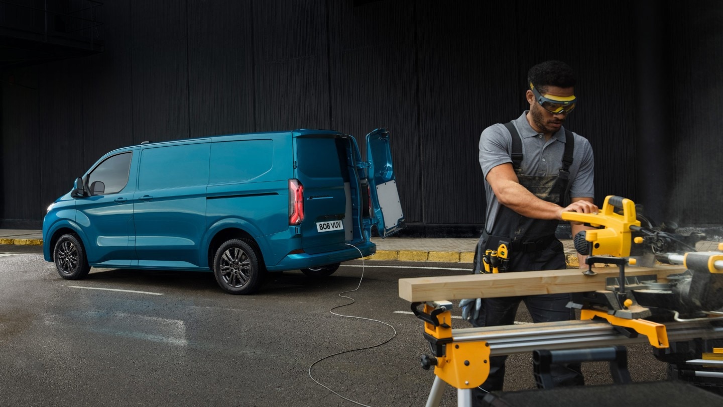 Ford E-Transit Custom in Blau. Dreiviertelansicht mit geöffneter Hecktür, Mann schneidet Holzbretter.