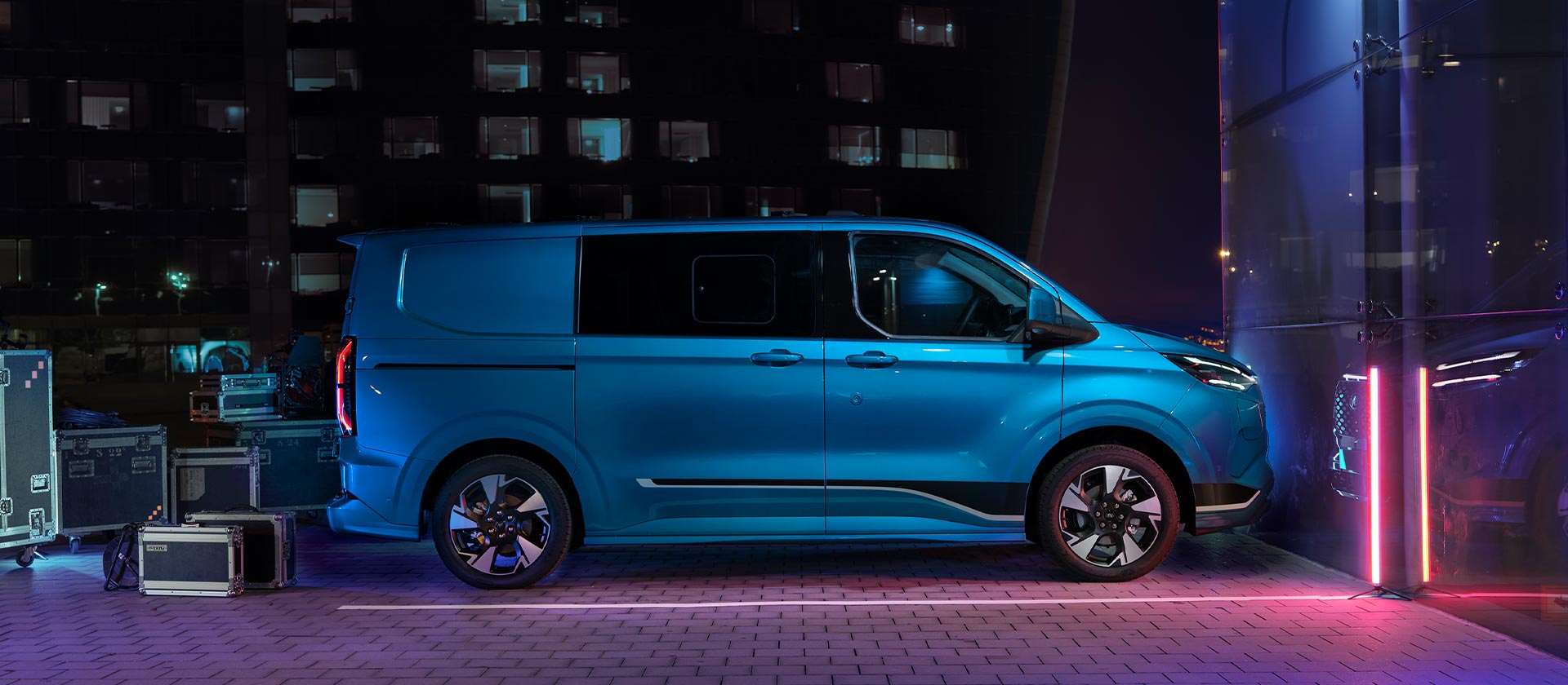 Ford E-Transit Custom in Blau. Seitenansicht, auf einem Parkplatz bei Nacht mit Kisten vor der Hecktür.