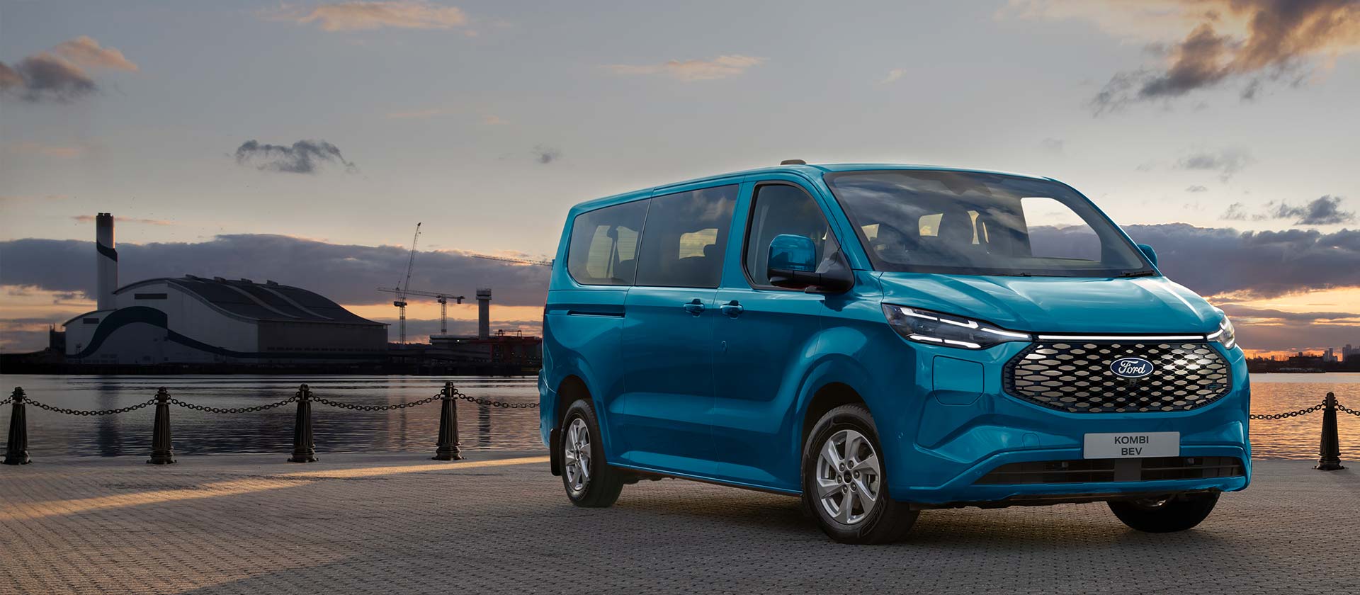 Ford E-Transit Custom Kombi in Blau. Dreiviertelansicht, parkt vor einem Hafen bei Sonnenuntergang.