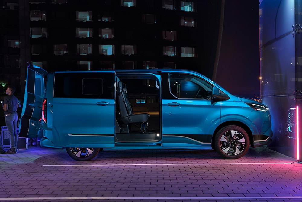 Ford E-Transit Custom in Blau. Seitenansicht, Person beladet den Transporter bei Nacht.