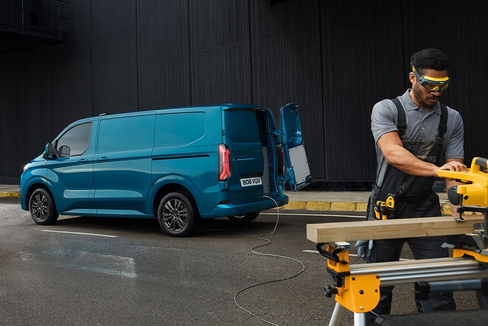 Ford Transit Custom in Blau. Dreiviertelansicht mit geöffneter Hecktür. Person schneidet Holzbretter.
