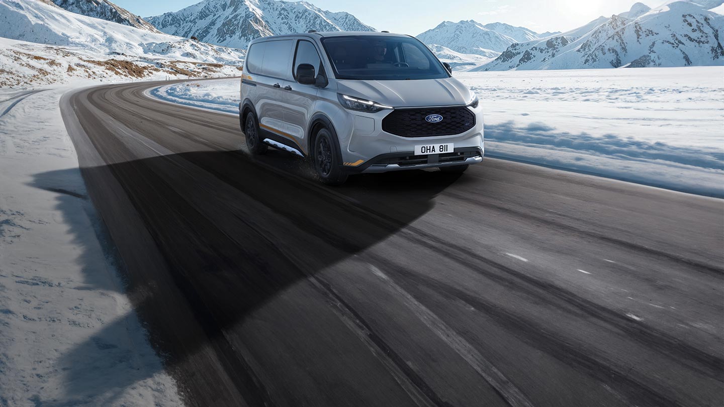 Ford E-Transit Custom AWD in Silber. Dreiviertelansicht, fährt auf einer Landstraße mit verschneiter Landschaft.