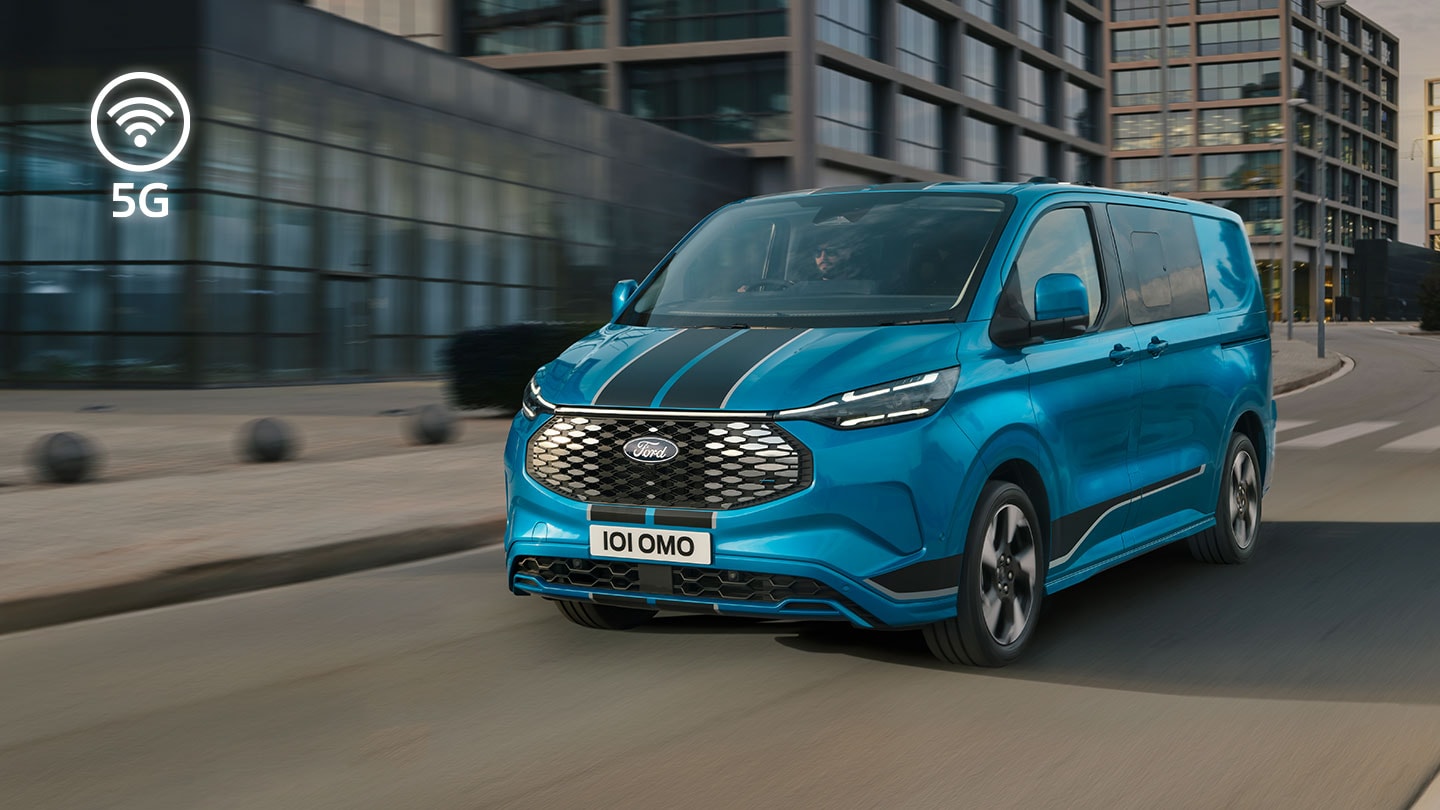 Ford E-Transit Custom in Blau. Dreiviertelansicht, fährt auf einer Straße neben Glasgebäuden. In der linken Ecke ist ein 5G-Logo zu sehen.