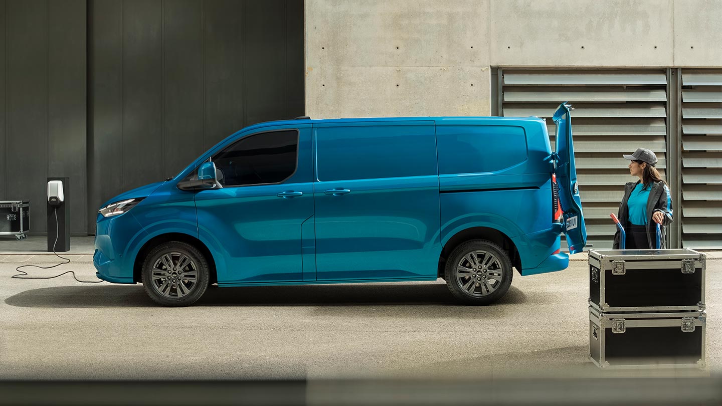 Ford E-Transit Custom Einzelkabine in Blau. Seitenansicht, an Ladestation angeschlossen. Eine Frau beladet währenddessen den Laderaum.