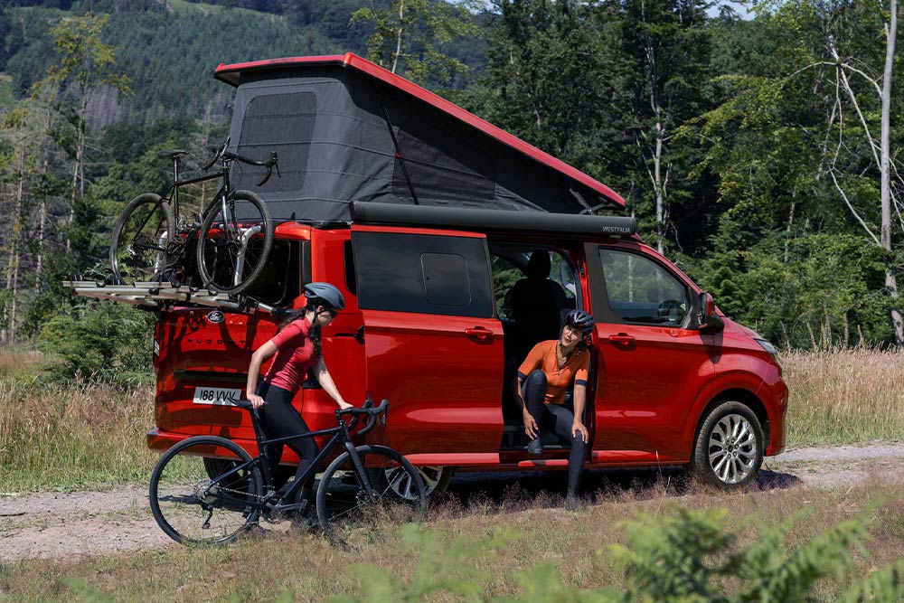 Ford Nugget in Rot. Heckansicht, mit geöffnetem Aufstelldach, Heckklappe, Fahrrädern und zwei Personen.