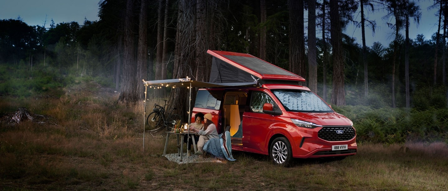 Ford Nugget in Rot. Dreiviertelansicht mit Aufstelldach, parkt in einem Wald 