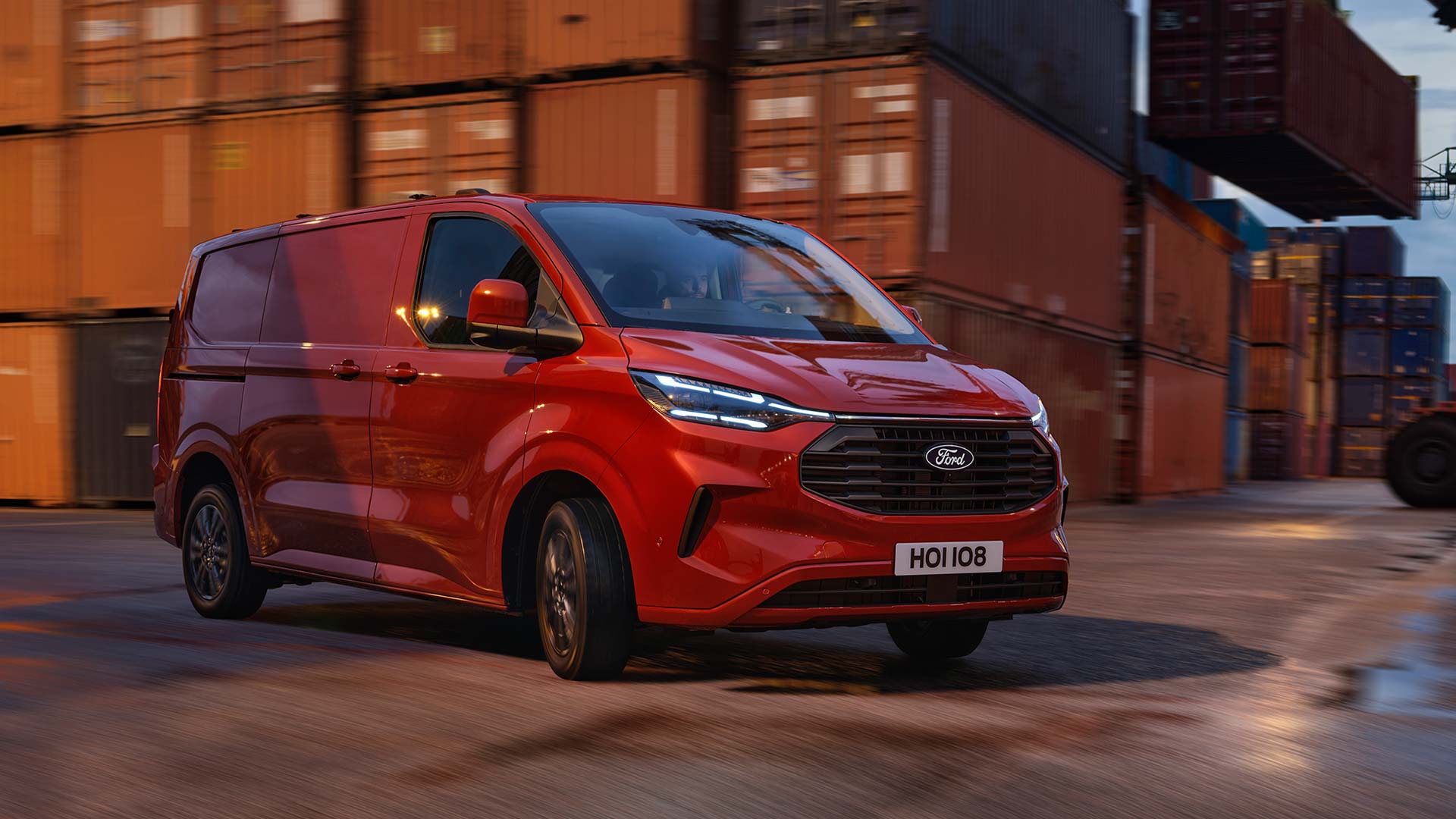 Ford Transit Custom in Rot. Dreiviertelansicht, fährt an gestapelten Containern vorbei.