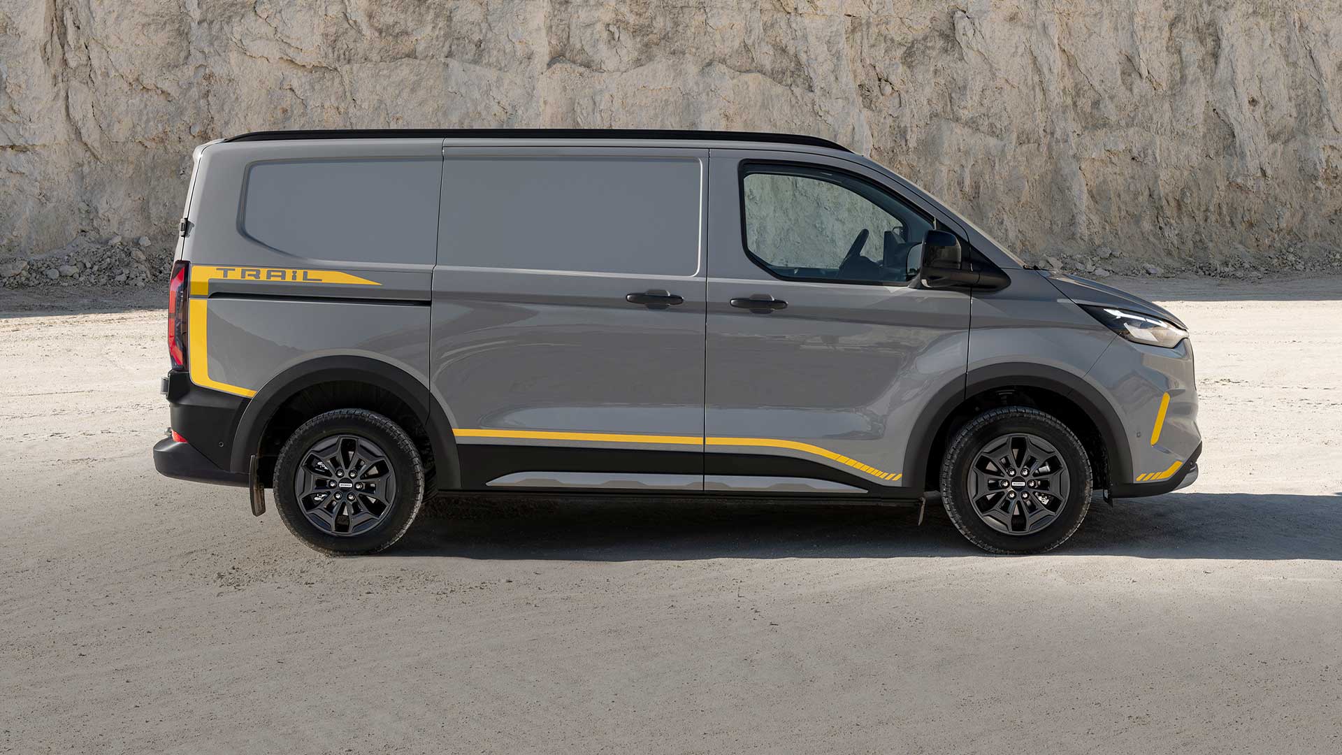 Ford Transit Custom Trail in Grau. Seitenansicht, parkt vor einer Felsmauer.