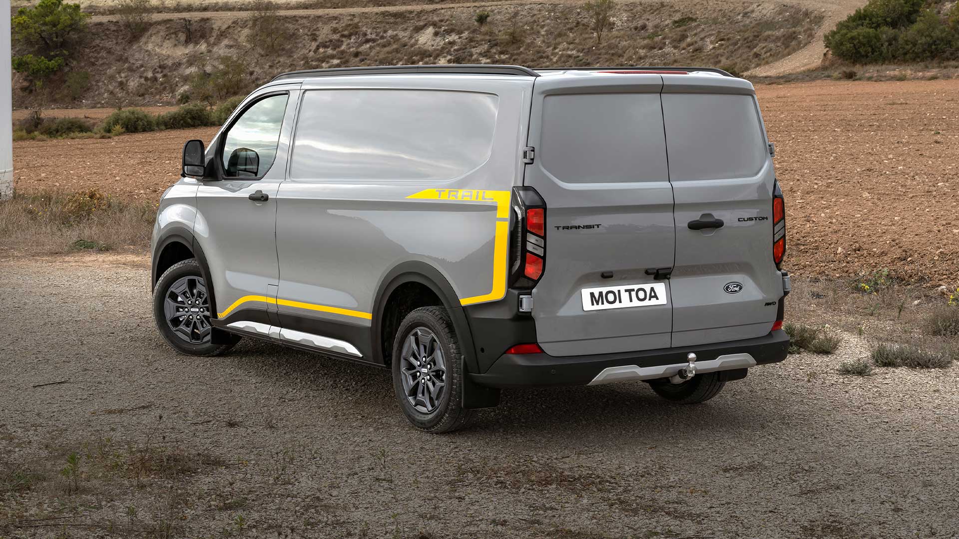 Ford Transit Custom Trail in Grau. Dreiviertelansicht vom Heck, parkt in einer trockenen Felslandschaft.