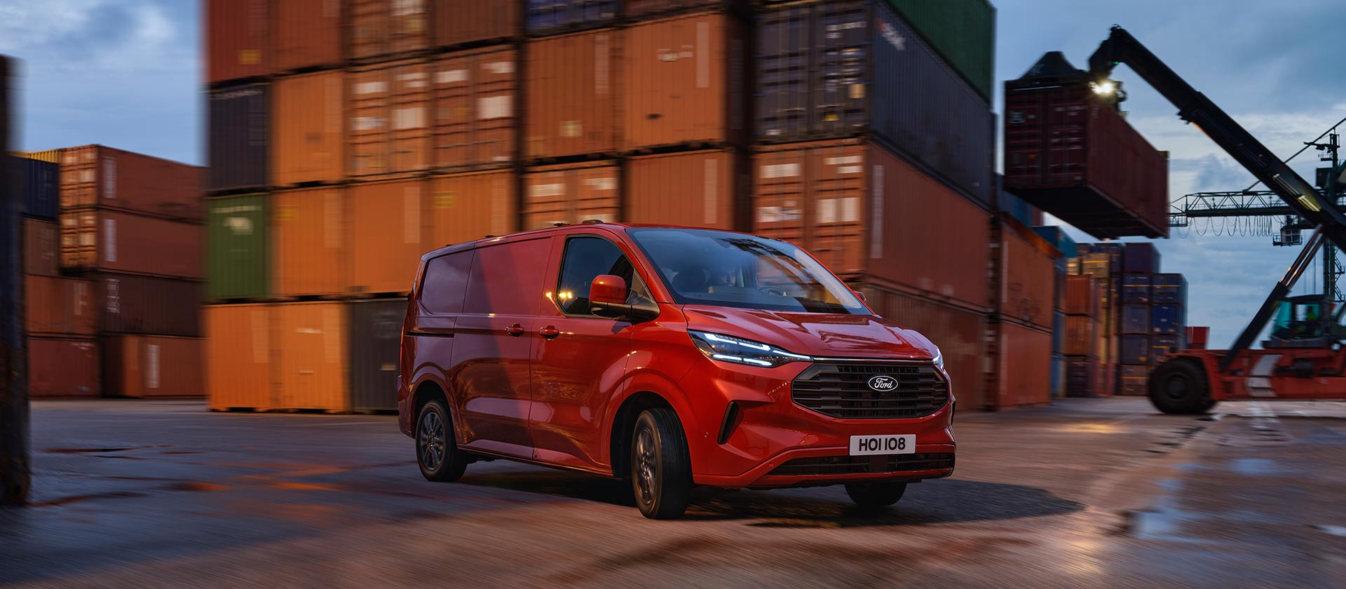 Ford Transit Custom in Rot. Dreiviertelansicht, fahrend auf einem Industriehafen mit Containern im Hintergrund.