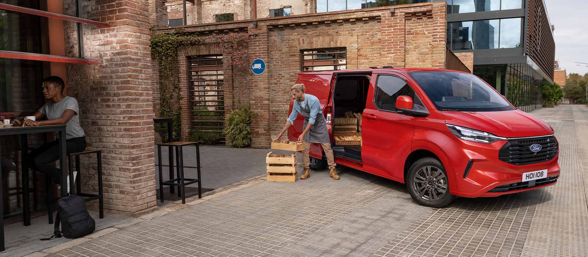 Ford Transit Custom Kastenwagen Einzelkabine in Rot. Dreiviertelansicht vor einem Backsteingebäude mit einem Mann der Ware aus dem Fahrzeug ausladet.