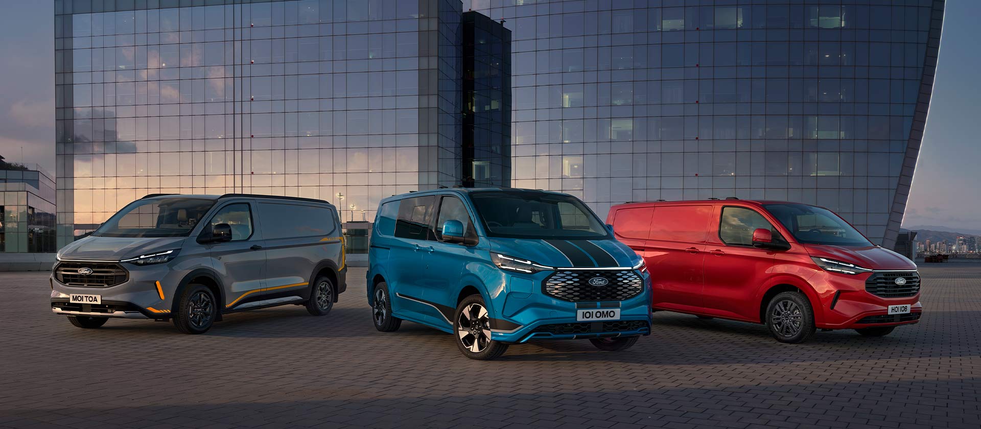 Drei nebeneinander geparkte Ford Transit Custom Fahrzeuge in Grau, Blau und Rot – parken vor modernen Glasgebäuden.
