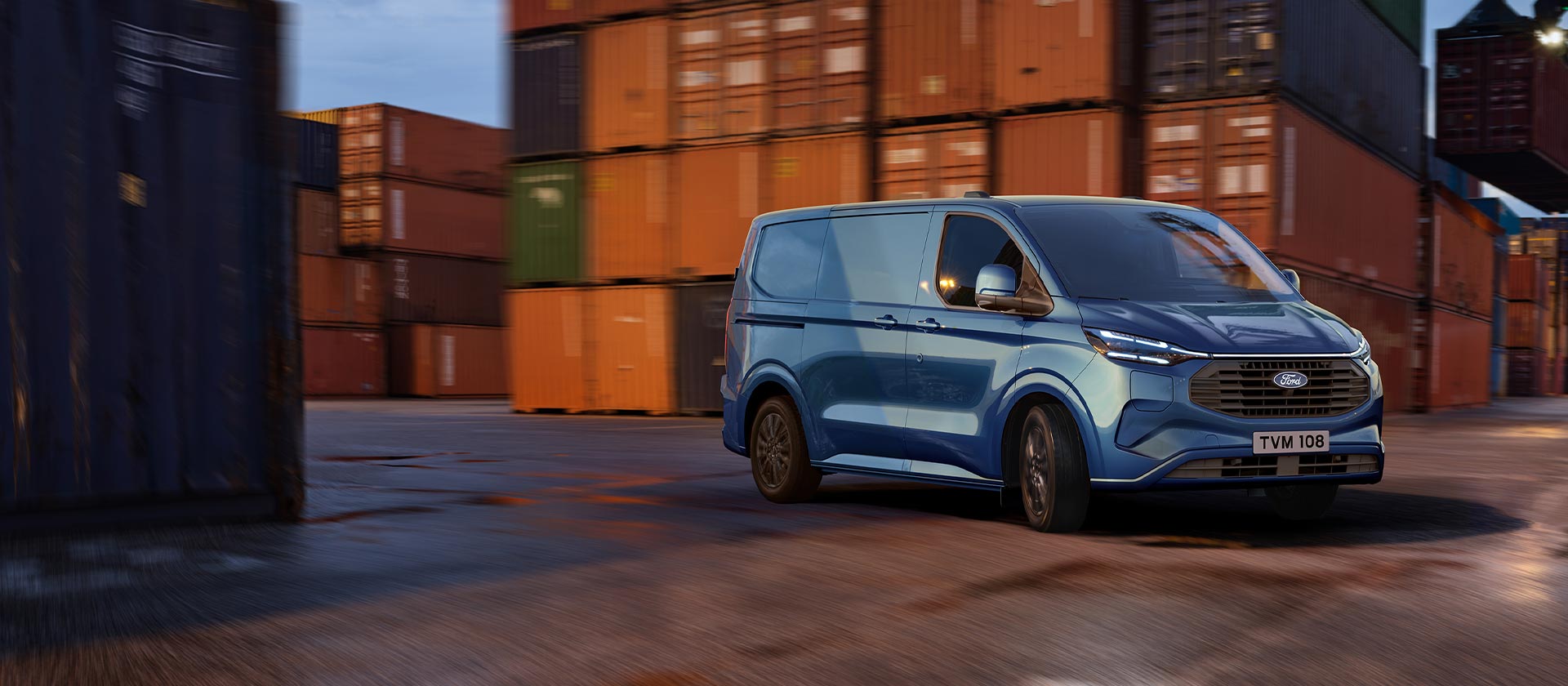 Ford Transit Custom Plug-in-Hybrid in Blau. Dreiviertelansicht, fährt an Containern vorbei.