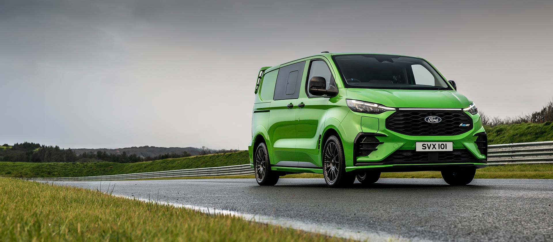 Ford Transit Custom MS-RT in Grün. Dreiviertelansicht, auf einer Rennstrecke mit Hügel im Hintergrund.