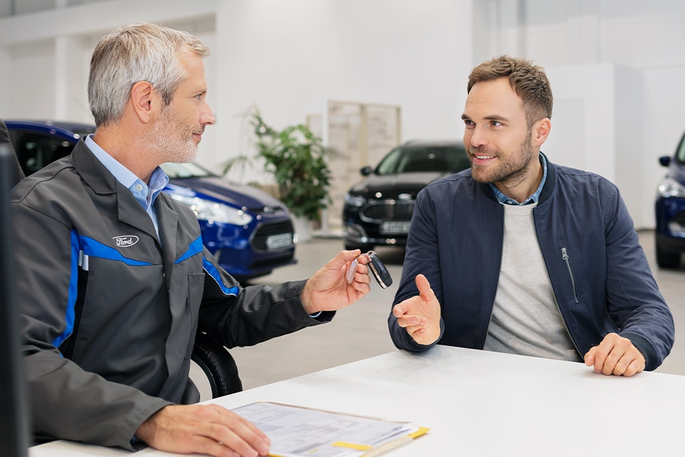 Zwei Personen unterhalten sich in einem Ford Autohaus. Im Hintergrund stehen Ford Fahrzeuge.