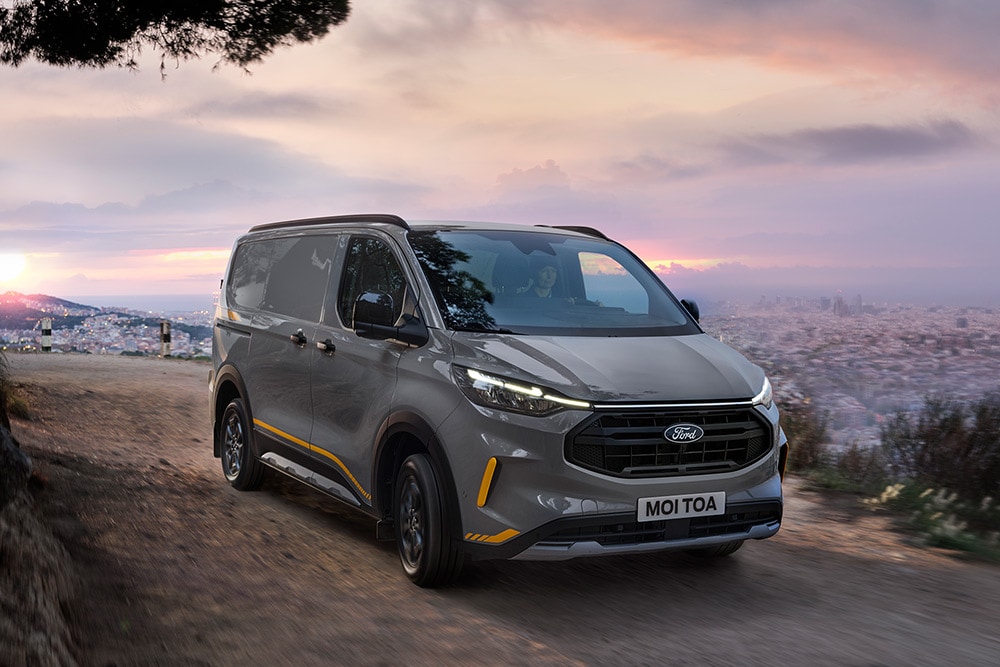 Ford Transit Custom Trail in Grau. Dreiviertelansicht, fahrend auf einer sandigen Straße bei Sonnenuntergang.