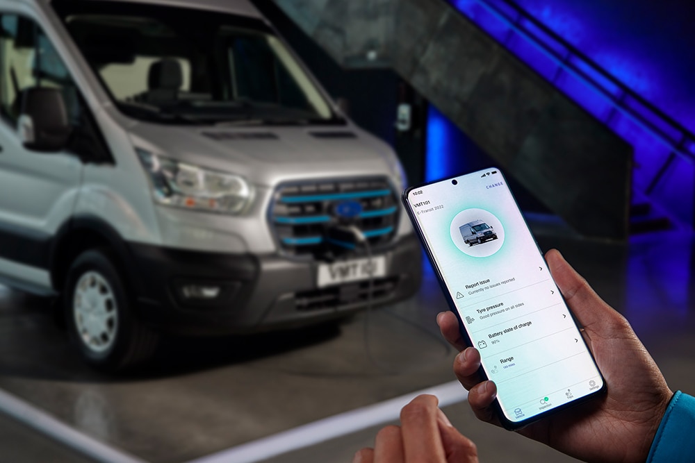 Eine Hand hält ein Smartphone mit geöffneter FordPass App. Im Hintergrund ist ein Ford Transit an einer Ladestation angeschlossen.
