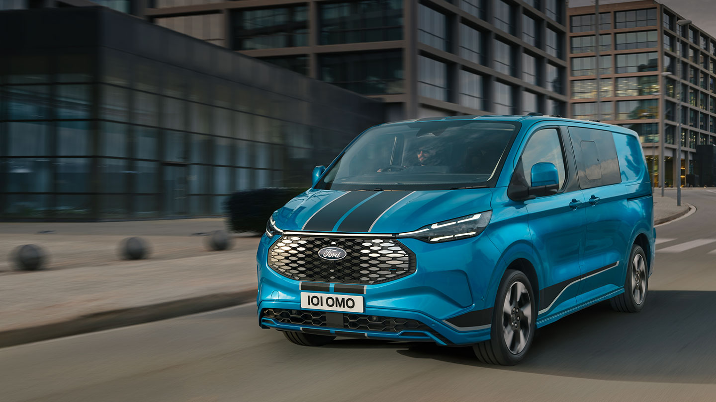 Ford E-Transit Custom in Blau. Dreiviertelansicht, fährt durch eine Innenstad mit mehreren Glasgebäuden.