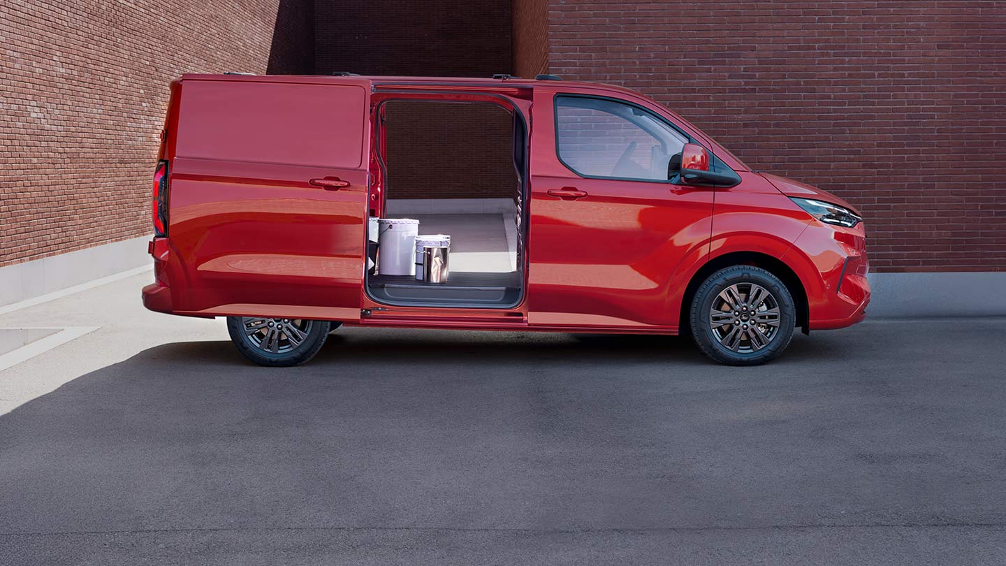 Ford Transit Custom Limited in Rot. Seitenansicht, stehend vor einer Ziegelsteinmauer mit geöffneter seitlicher Schiebetür.
