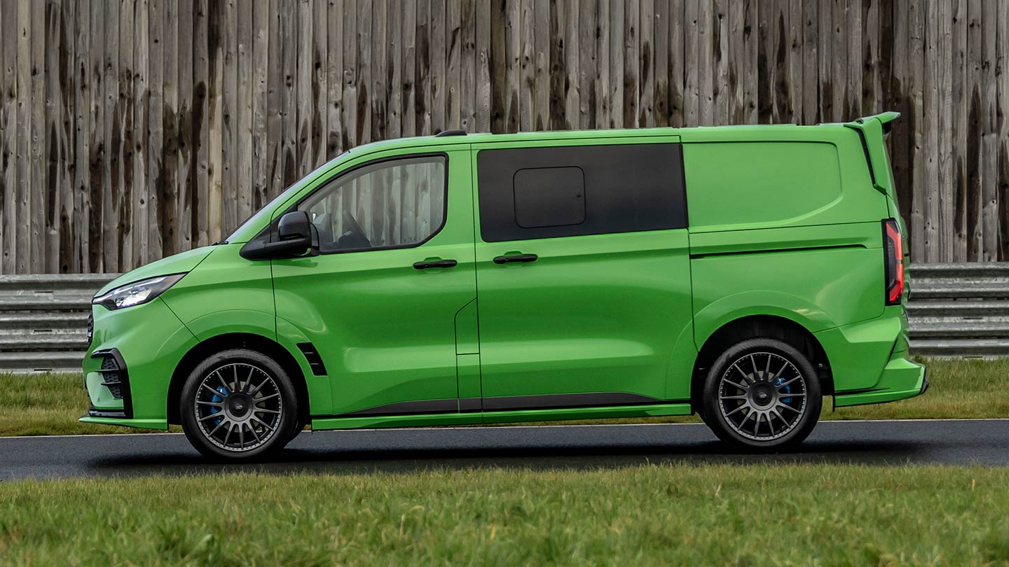Ford Transit Custom MS-RT in Grün. Seitenansicht, parkt vor einer Holzwand.