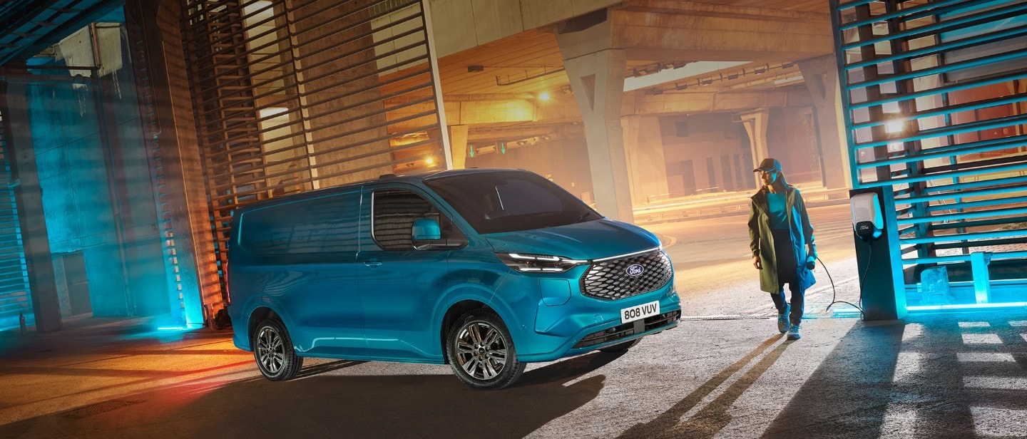 Ford E-Transit Custom in Blau. Dreiviertelansicht, stehend in einer Parkgarage neben einer Ladestation. Frau mit Ladekabel in der Hand nähert sich dem Fahrzeug.