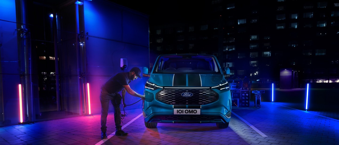 Ford E-Transit Custom in Blau. Frontansicht, Mann schließt Ladekabel an das Fahrzeug an.