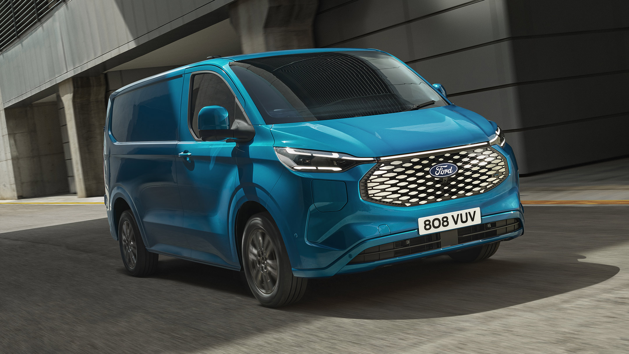 Ford E-Transit Custom in Blau. Dreiviertelansicht, fahrend auf einer Straße. Gebäude im Hintergrund.