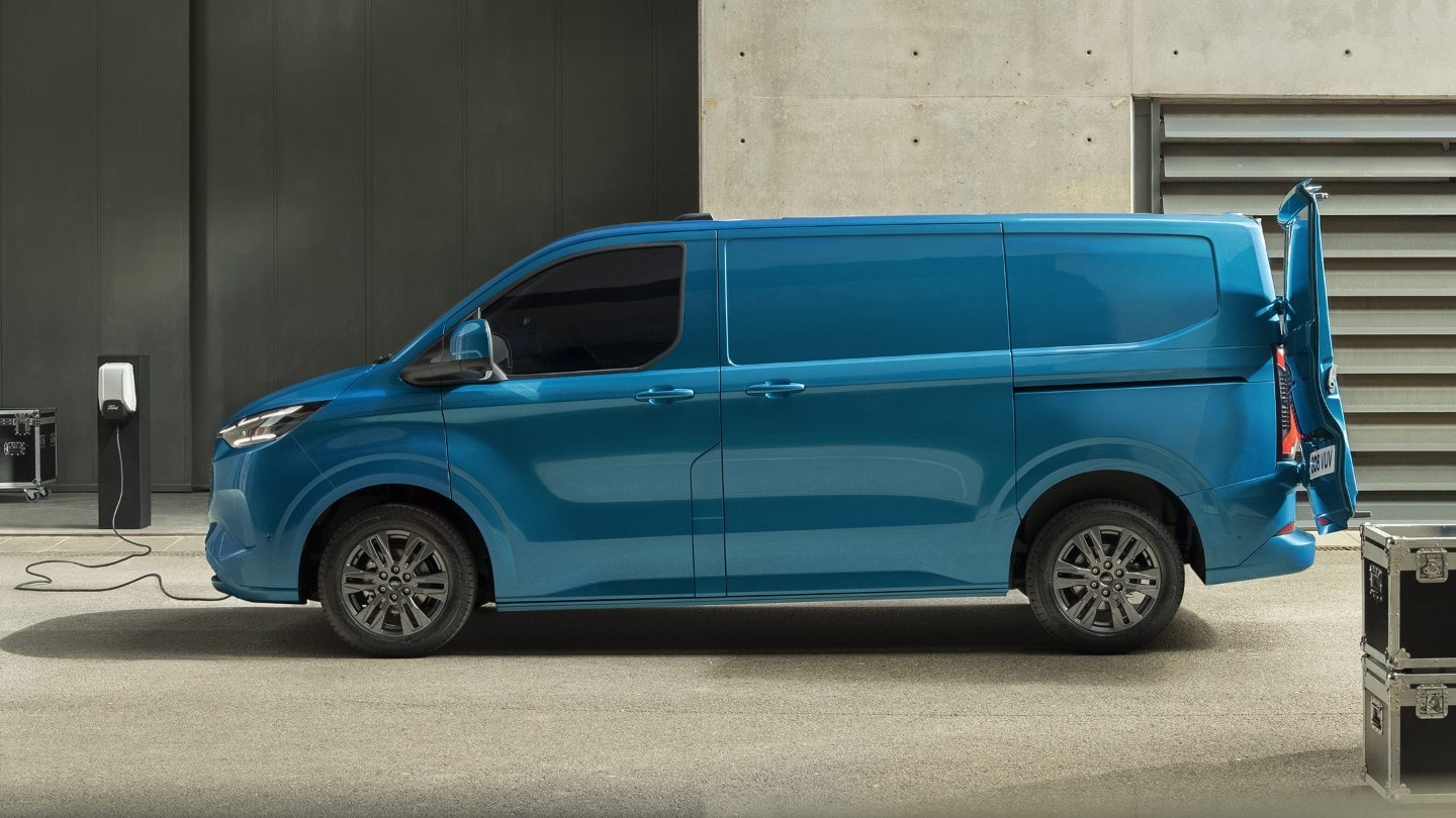 Ford E-Transit Custom in Blau. Seitenansicht, stehend vor einem Gebäude an einer Ladestation angeschlossen.