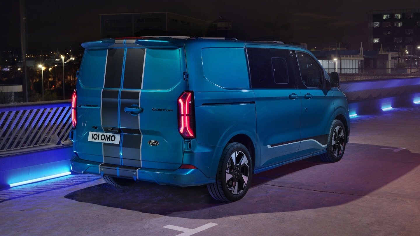 Ford E-Transit Custom in Blau. Dreiviertelansicht, fahrend über eine Brücke bei Nacht.