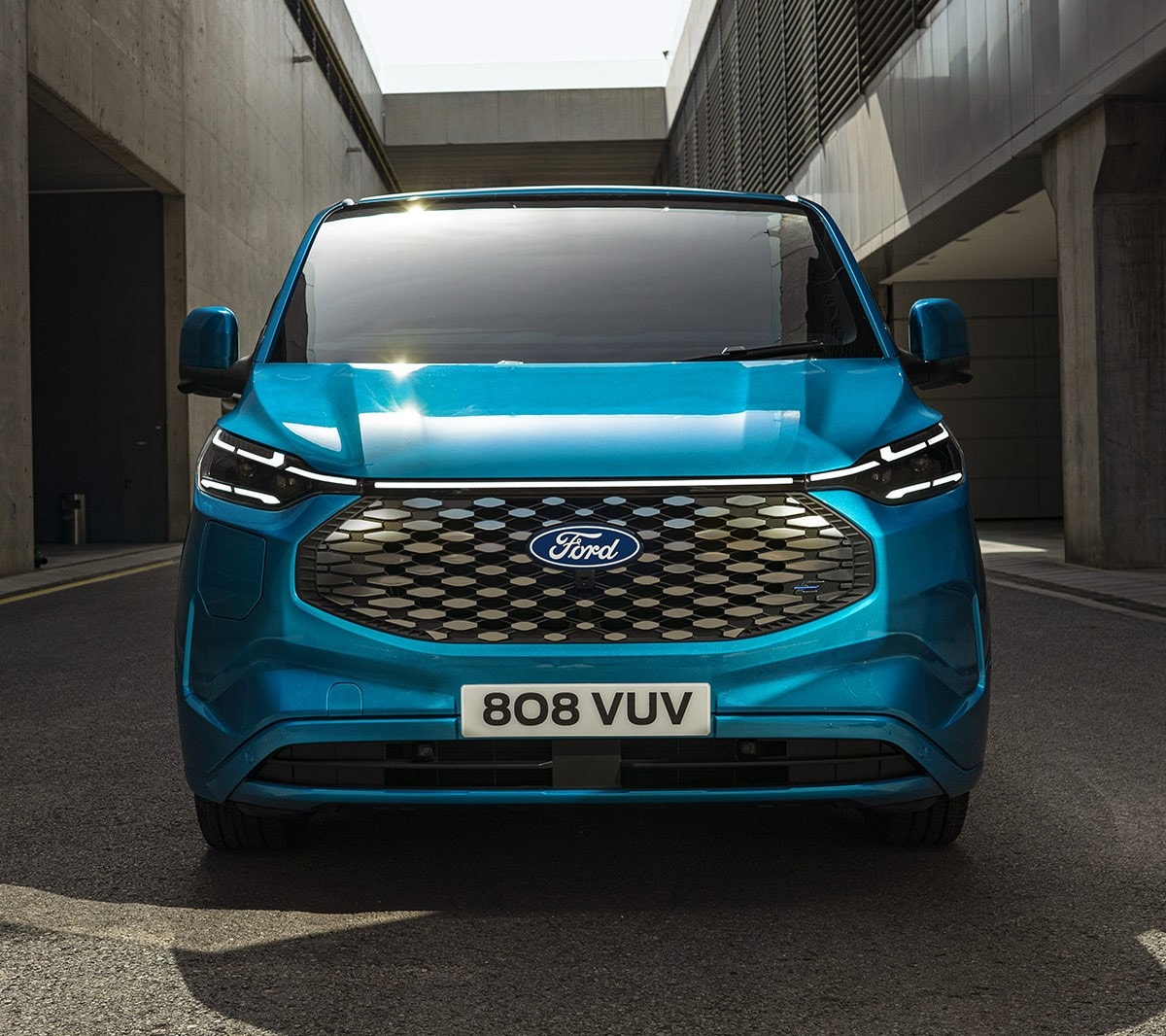 Ford E-Transit Custom in Blau. Frontansicht, fahrend auf einer Straße. Gebäude im Hintergrund.