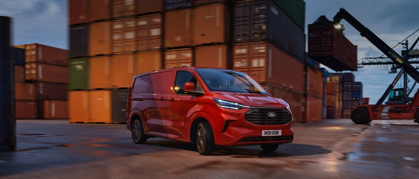 Ford Transit Custom Limited in Rot. Dreiviertelansicht, fahrend auf einer Straße mit Containern im Hintergrund.