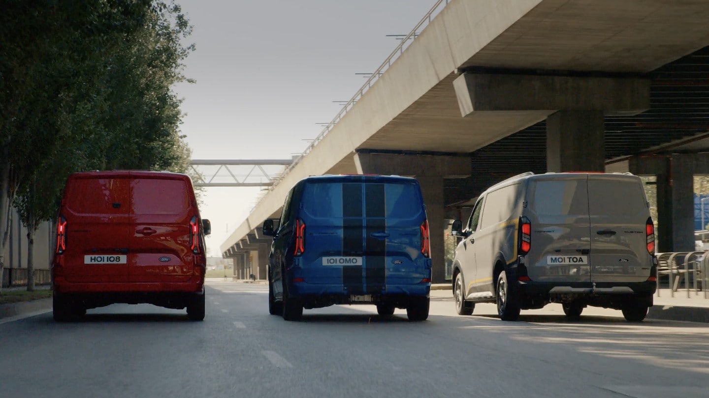 Ford Transit Custom Limited in Rot, Ford Transit Custom in Blau und Ford Transit Custom Trail in Grau. Heckansicht, fahren nebeneinander auf einer Straße.